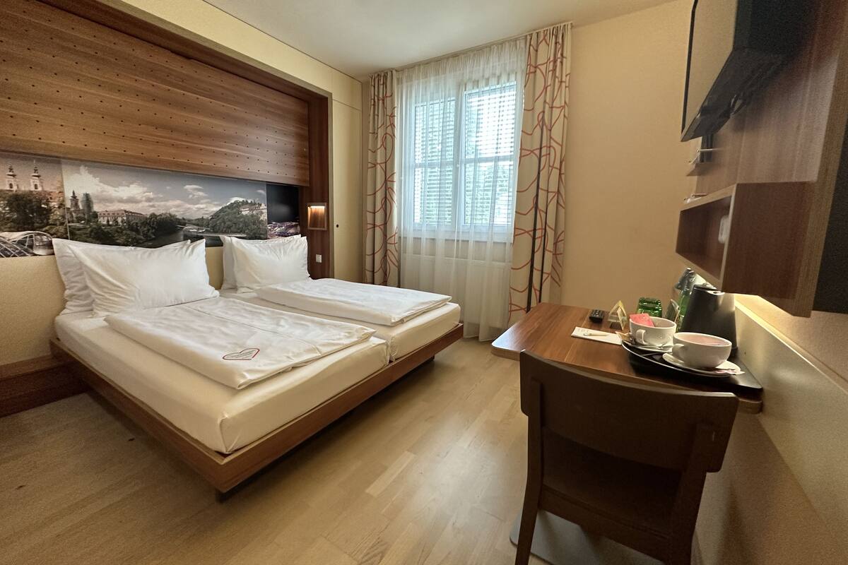 A hotel room with two single beds, a wooden headboard, a table with a cup and saucer, and a view of a cityscape through the window.
