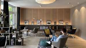 A modern lounge area with a high ceiling, recessed lighting, a geometric ceiling design, and a seating area with multiple chairs and a sofa.