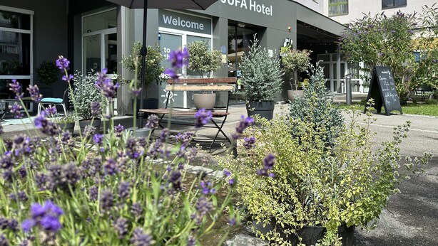 A hotel entrance with a 'Welcome' sign, outdoor seating, and potted plants.