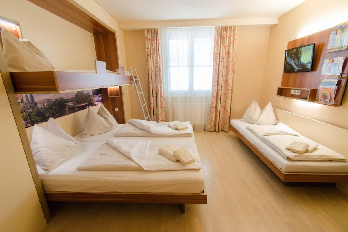 A hotel room with two single beds, a wooden bunk bed, a TV mounted on the wall, and a window with curtains.