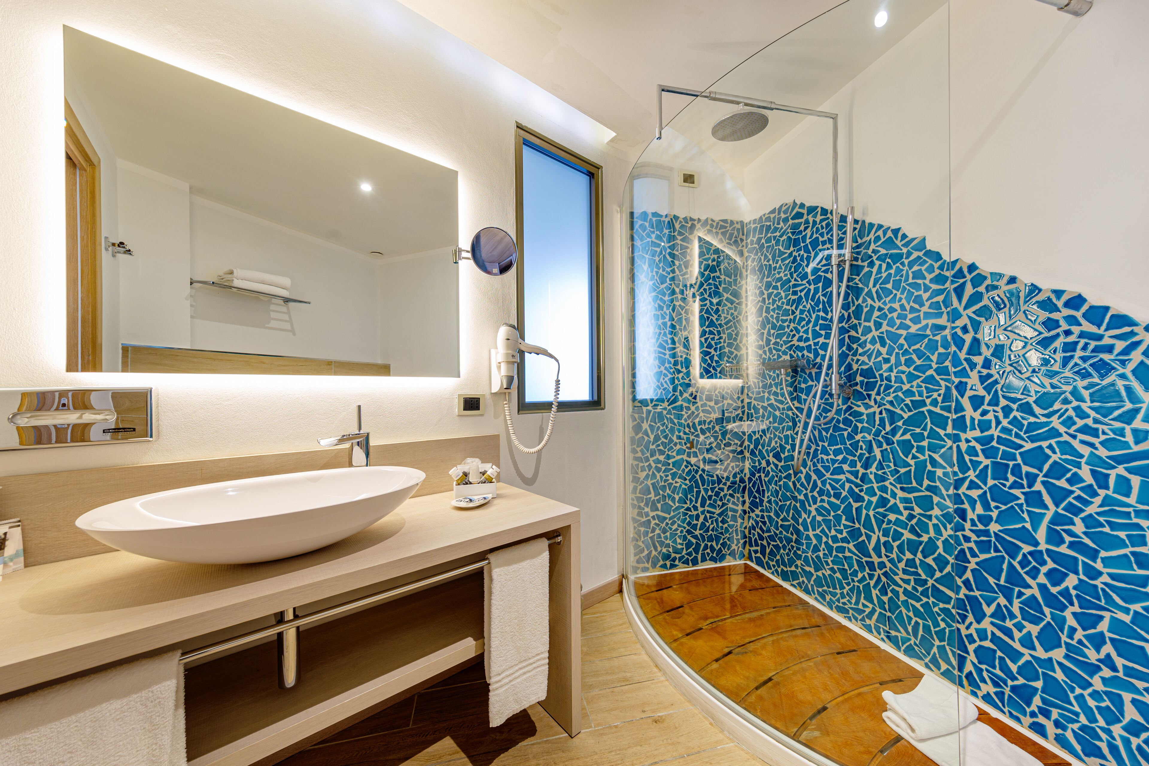 Un bany modern amb un gran mànega blanca, un mirall i una dutxa amb portes de vidre i mosaics blaus.