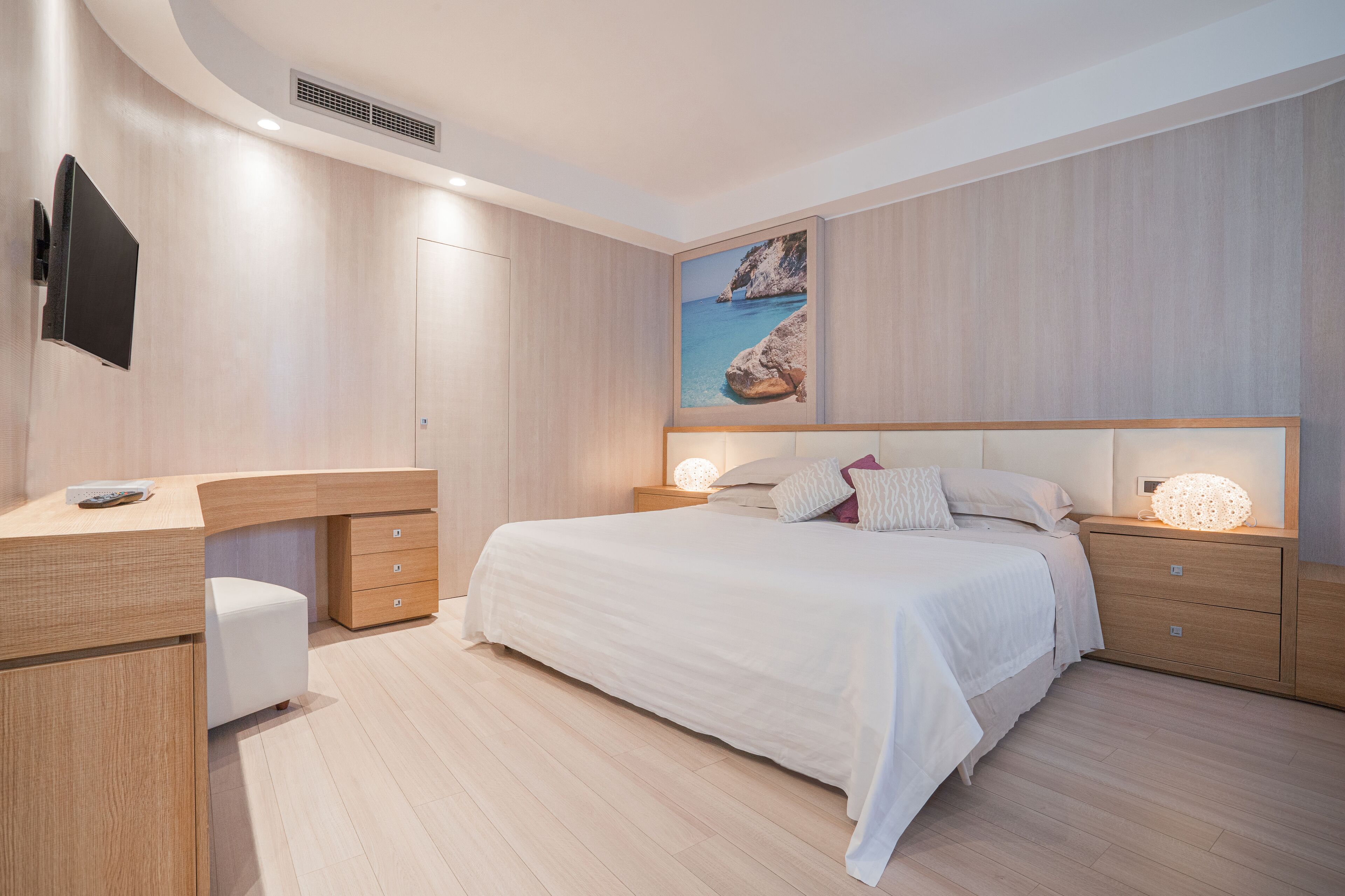 A modern bedroom with a wooden headboard, a flat-screen TV mounted on the wall, a bedside table with a lamp, and a large painting of a coastal scene.