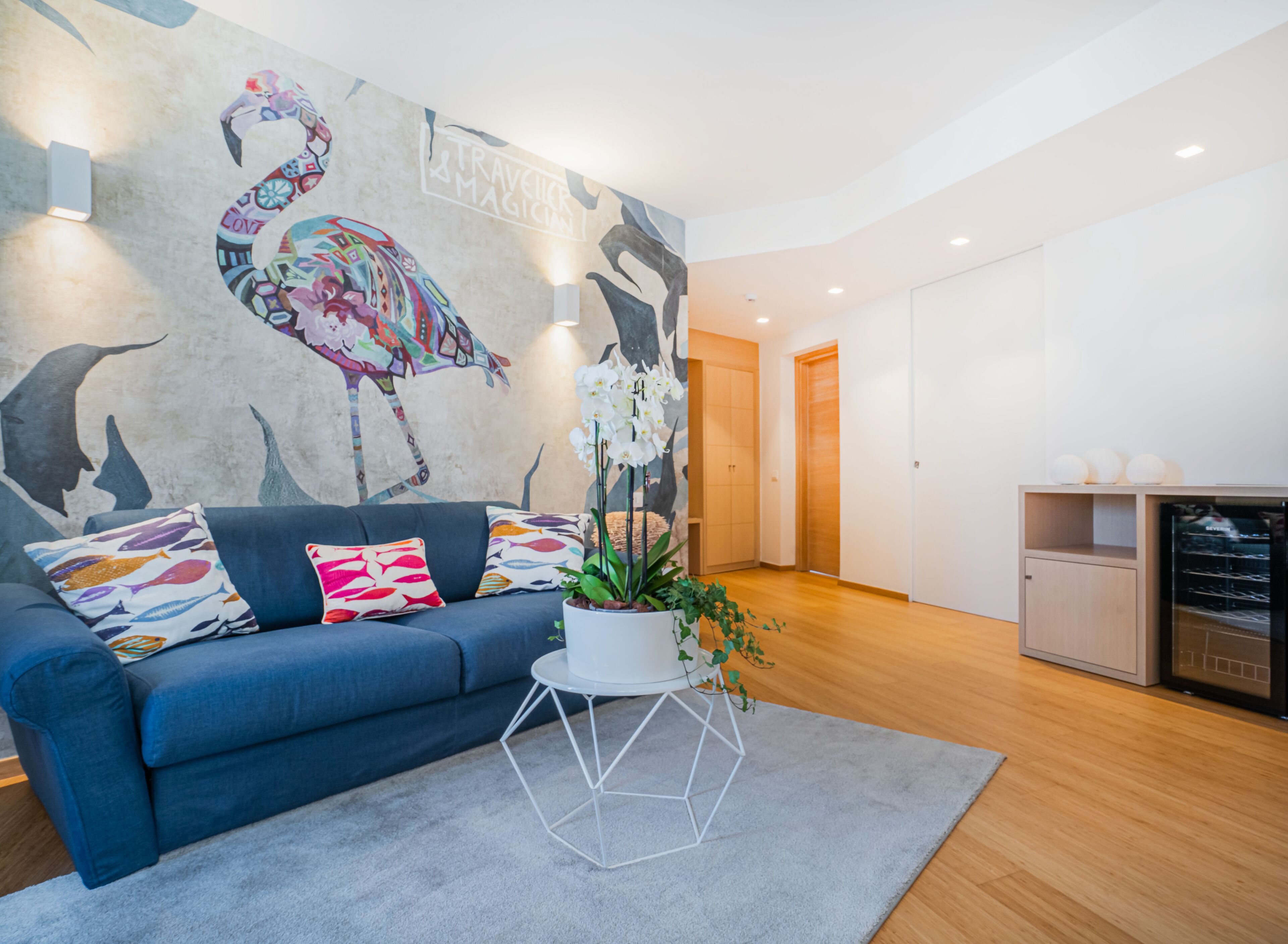 A modern living room with a blue sofa, colorful pillows, a white geometric coffee table, and a large wall mural.