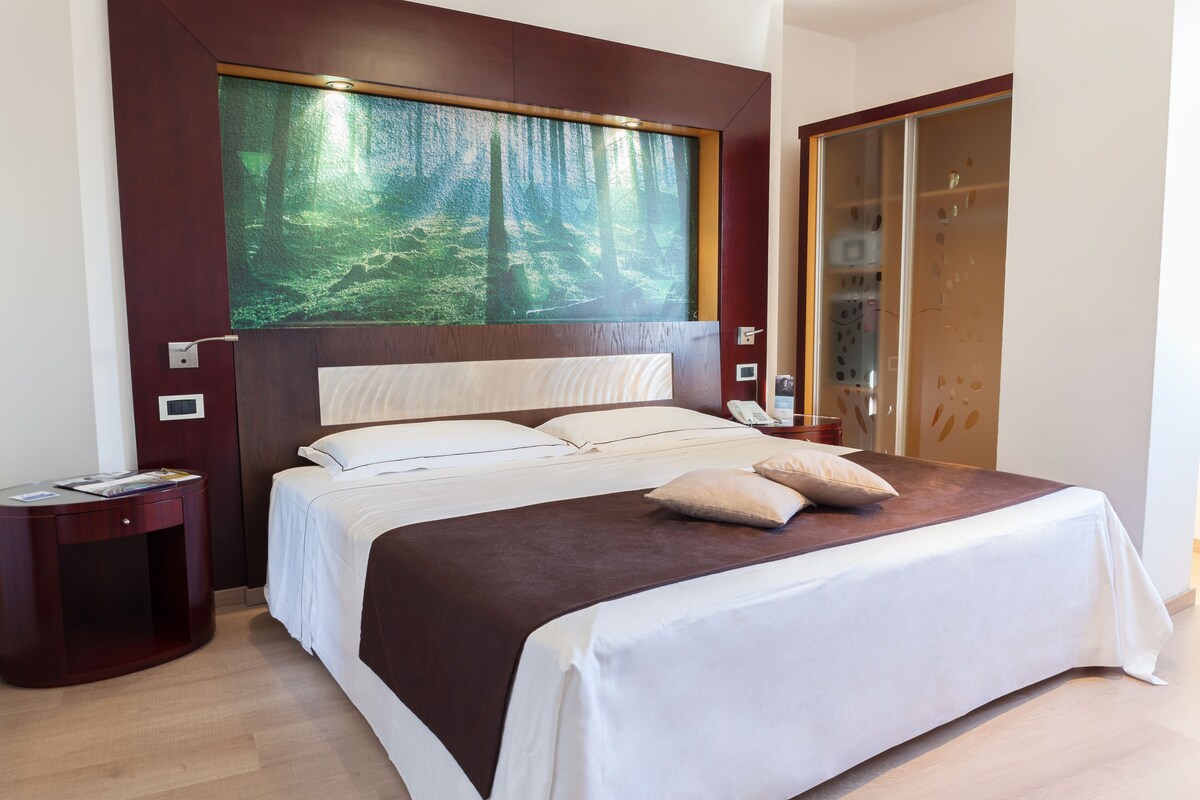Une chambre d’hôtel avec un grand lit, une table de chevet, un tableau représentant une forêt et une armoire en bois.