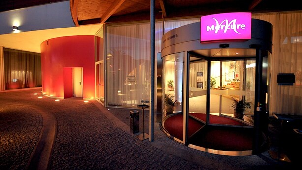 Une entrée d’hôtel moderne avec une porte en verre, une façade illuminée en rouge et un espace de réception bien visible à l’intérieur.