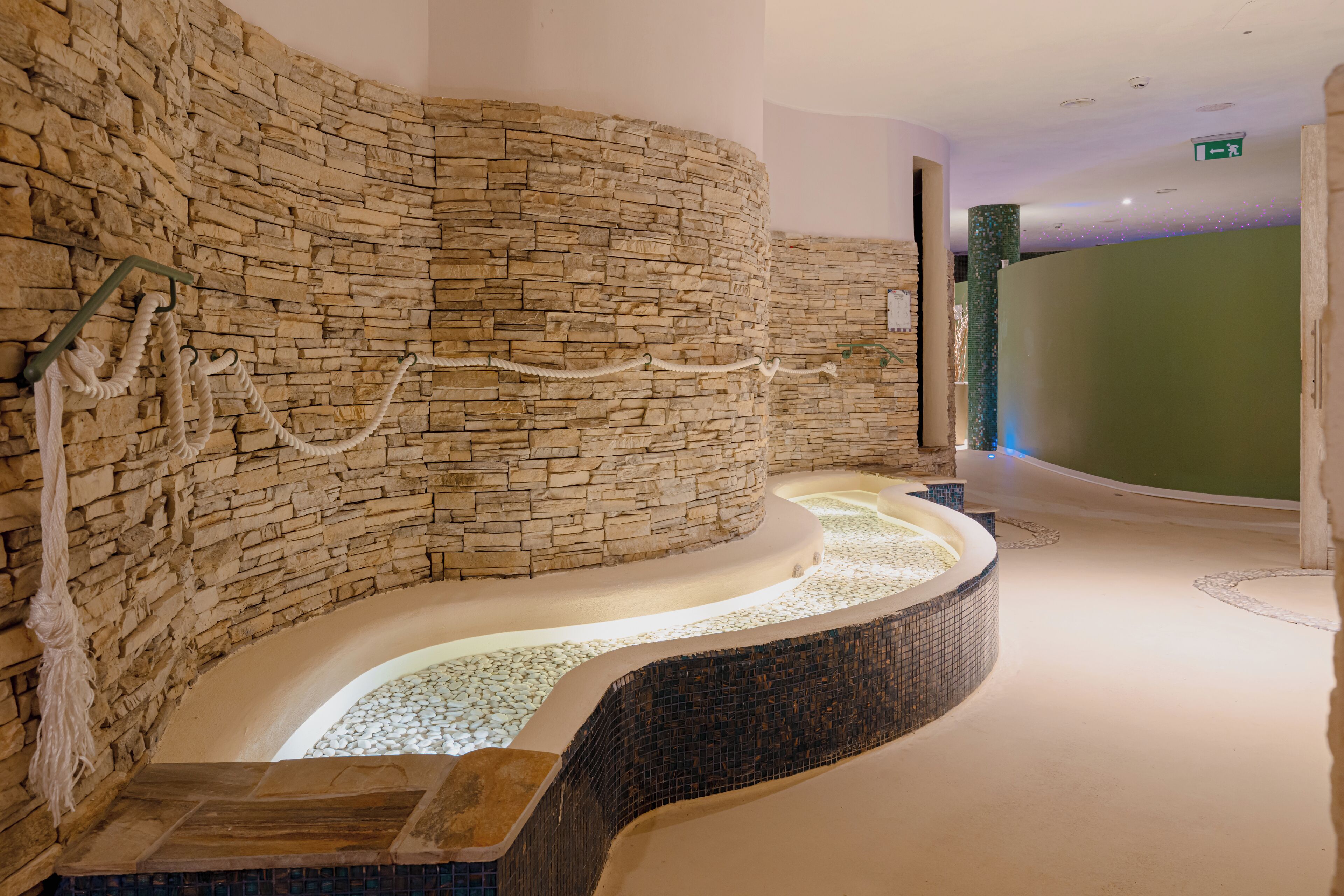 Un'area benessere con parete in pietra, elemento d'acqua curvo e una panca in legno.