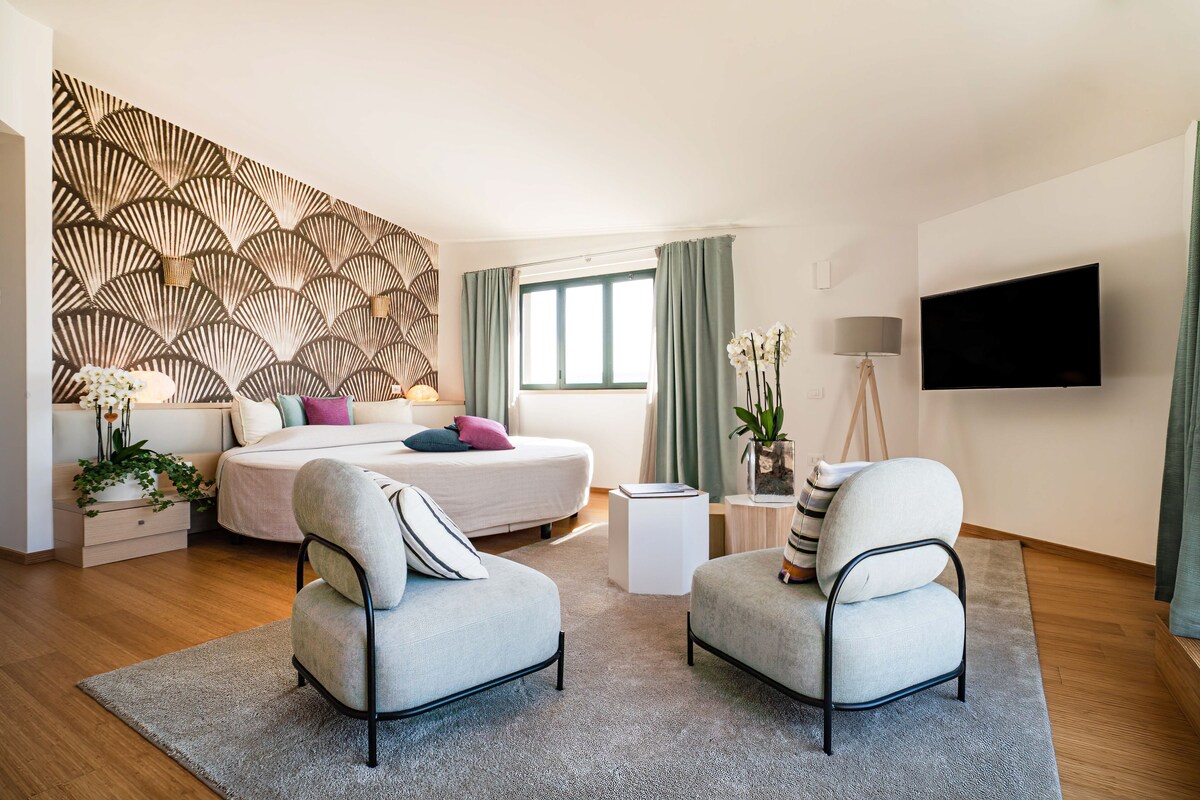 Une chambre d’hôtel moderne dotée d’un grand lit, de deux fauteuils, d’un téléviseur à écran plat et d’un panneau décoratif mural.