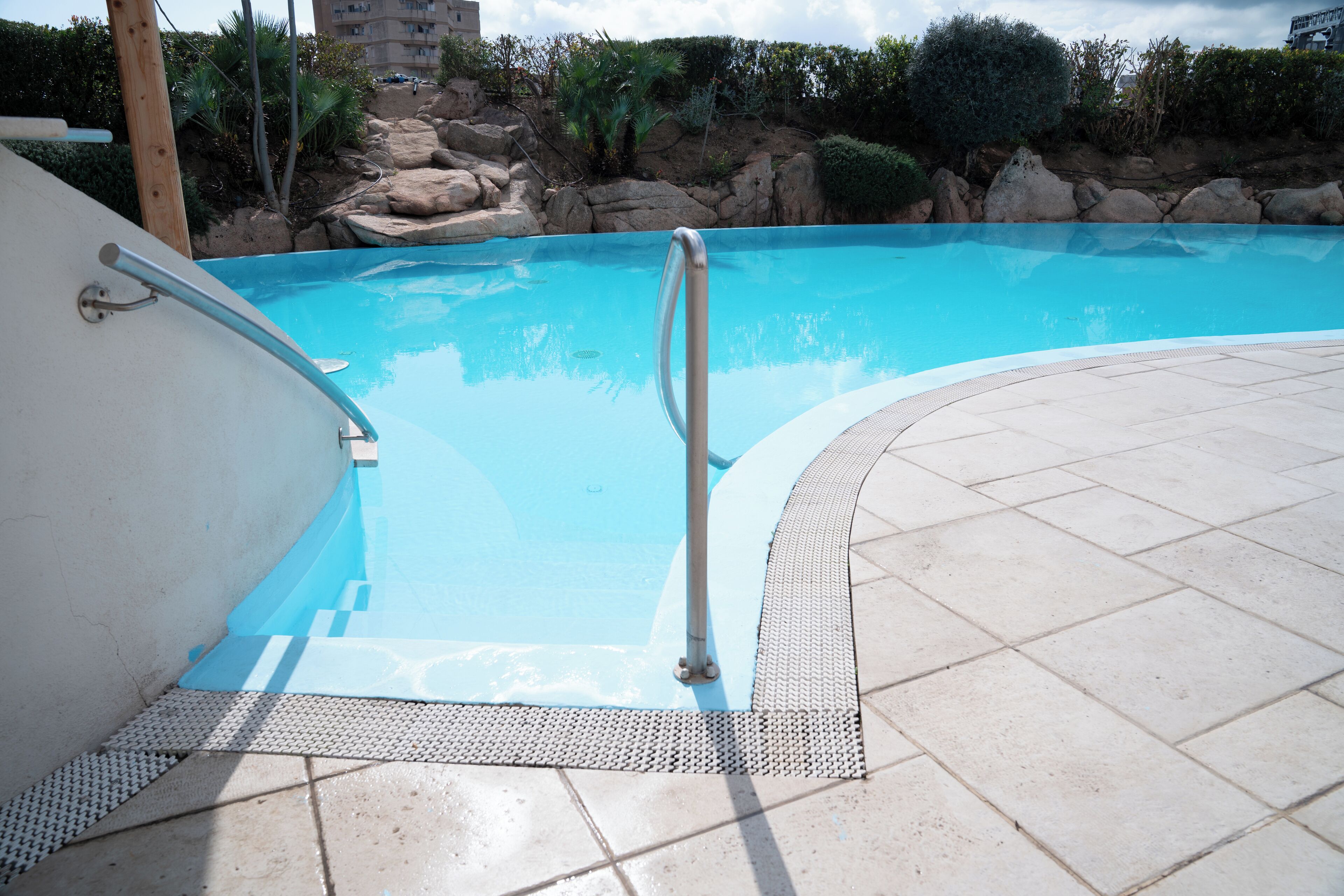 A swimming pool with a metal handrail, tiled deck, and a rocky landscape in the background.