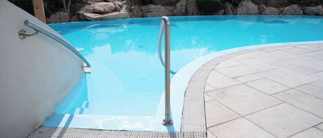 A swimming pool with a metal handrail, tiled deck, and a rocky landscape in the background.
