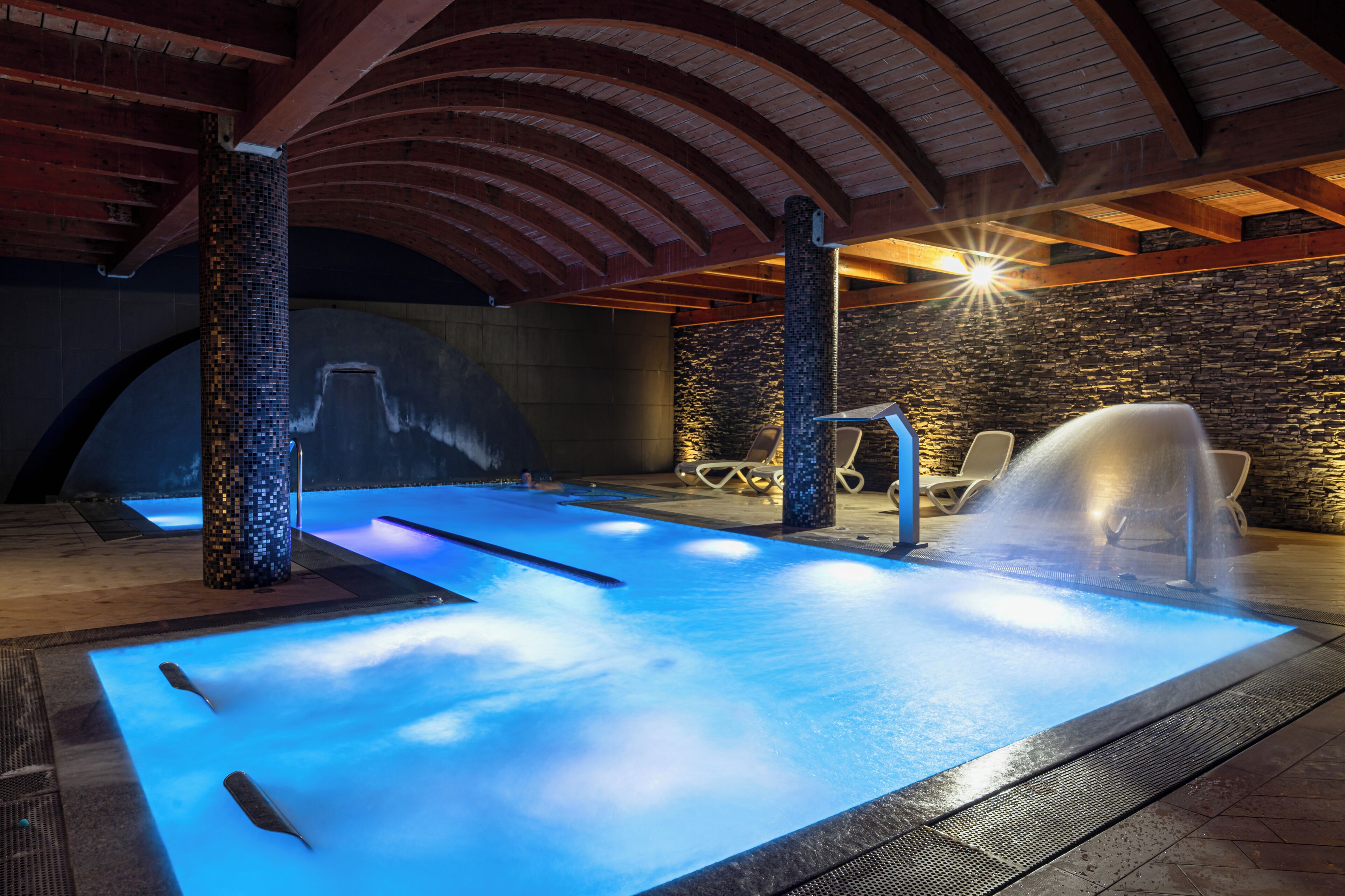 Una piscina interior con un elemento acuático iluminado en azul, paredes de piedra y vigas de madera.