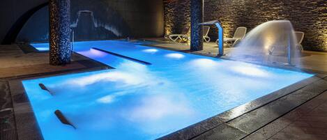 Una piscina interior con un elemento acuático iluminado en azul, paredes de piedra y vigas de madera.