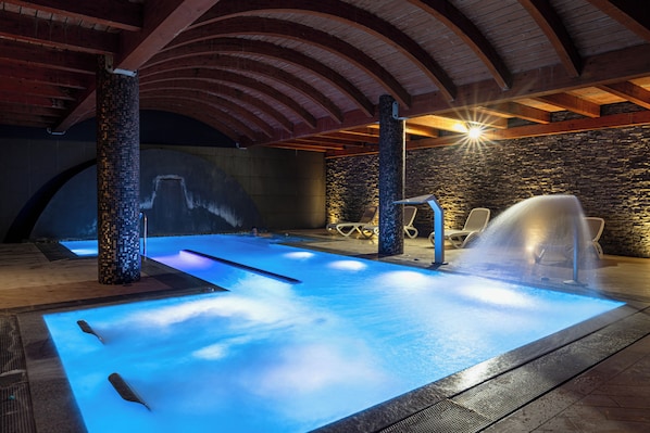 Una piscina interior con un elemento acuático iluminado en azul, paredes de piedra y vigas de madera.
