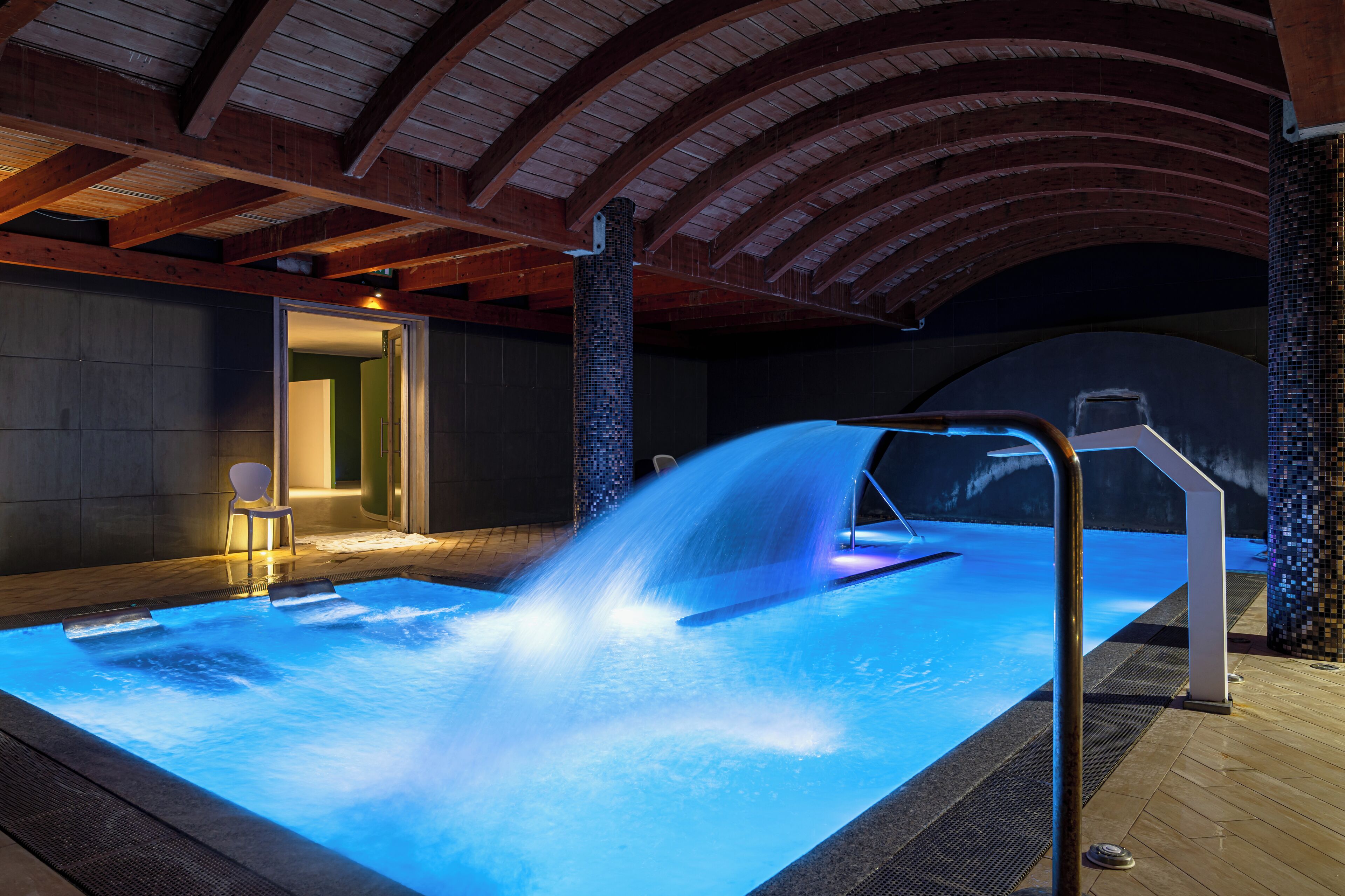 Una piscina interior climatizada con caída de agua, vigas de madera en el techo y pared de azulejos.