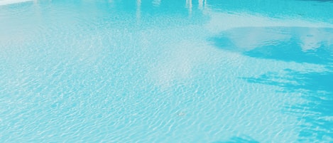 Una piscina con superficie de agua azul cristalina, una zona de estar cubierta y un edificio al fondo.