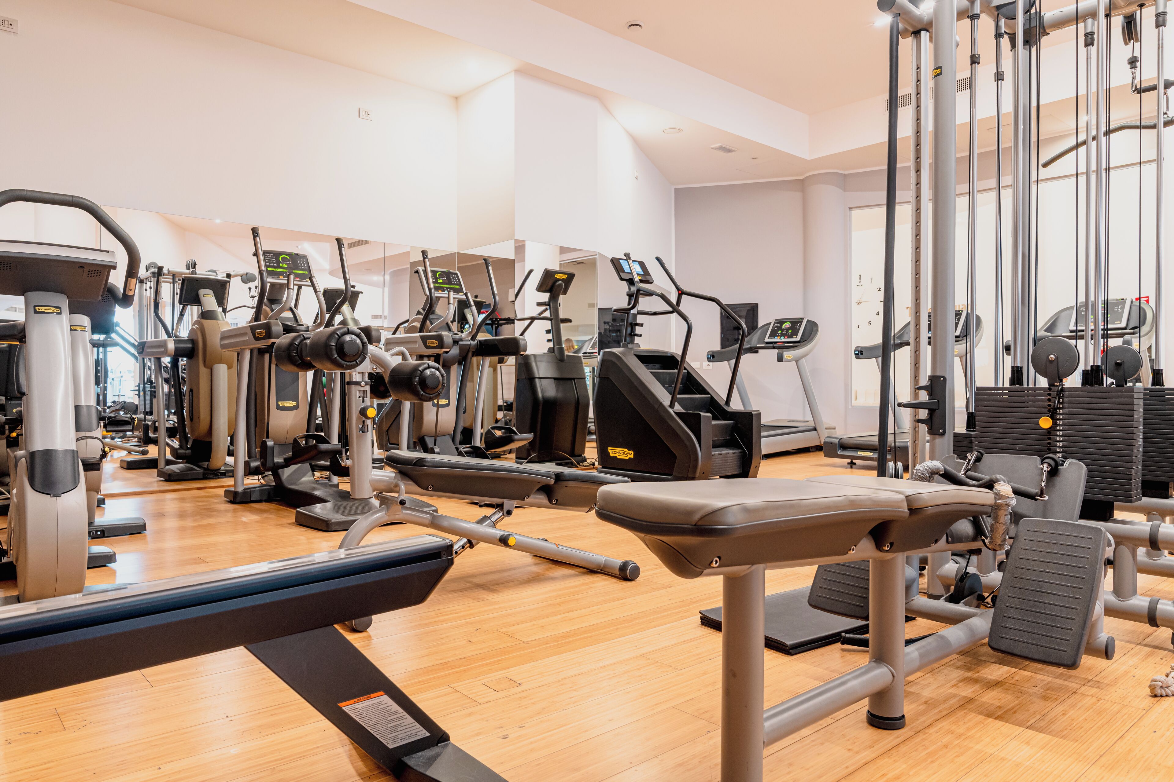 Un gimnasio con diferentes máquinas de ejercicio, piso de madera y un ventanal grande.