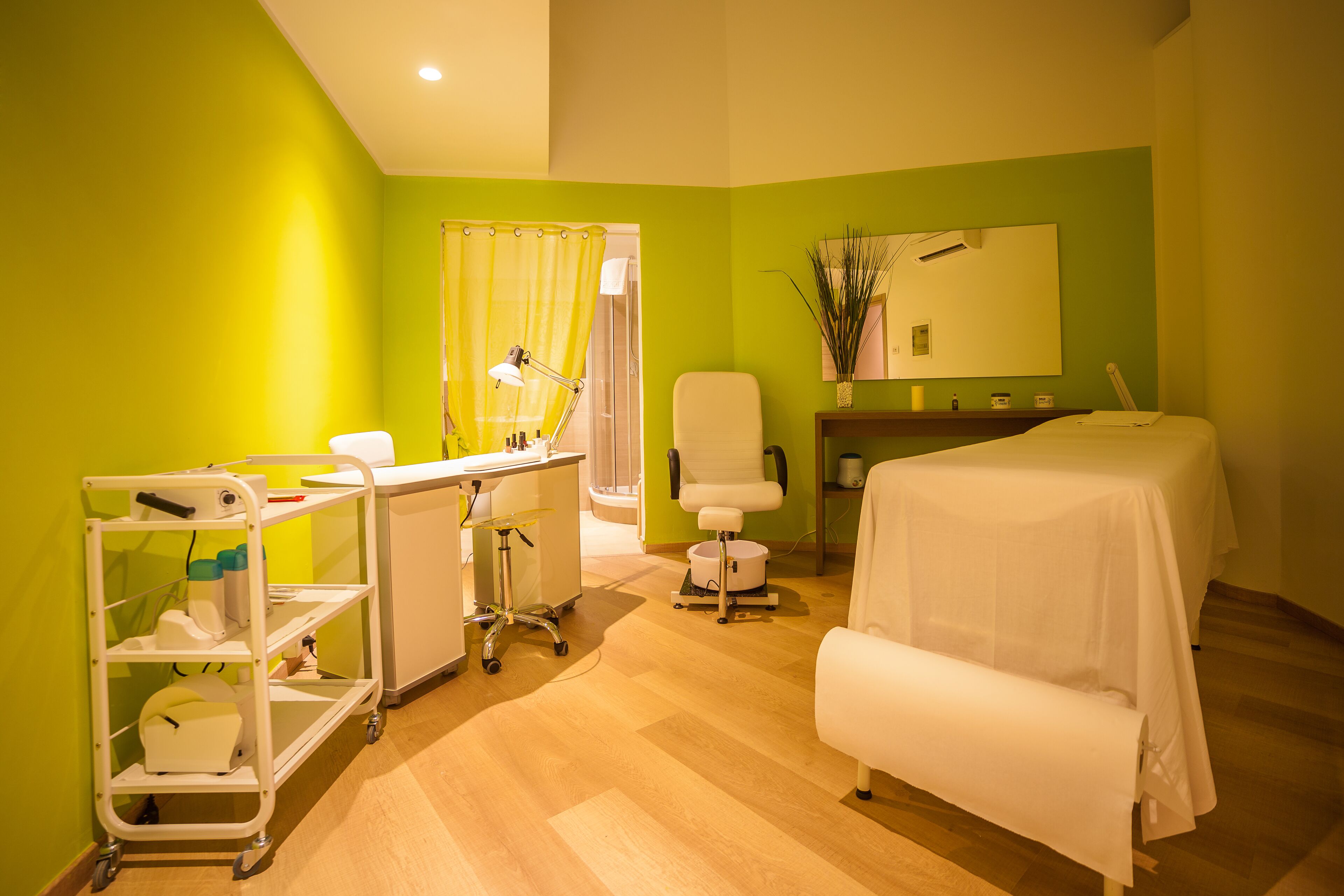 Quarto com parede verde vibrante, escrivaninha branca com um computador, uma cadeira, cama com cobertura branca e uma pequena mesa com espelho e plantas decorativas.