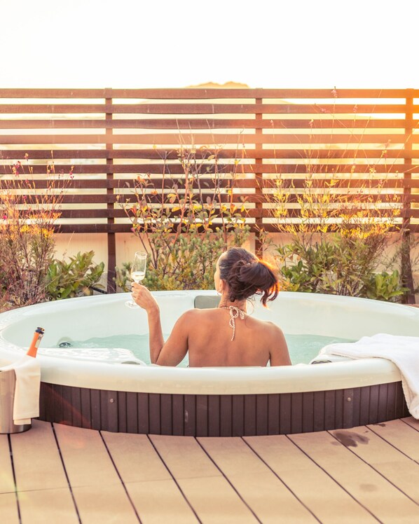 Une personne se détend dans un bain à remous, avec une terrasse en bois et des plantes en arrière-plan.