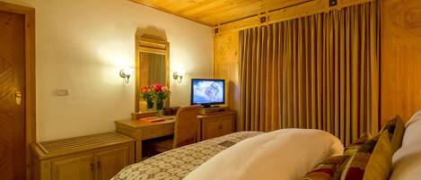 A hotel room with a wooden ceiling, a bed with patterned bedding, a desk with a television, a mirror, and a window with curtains.