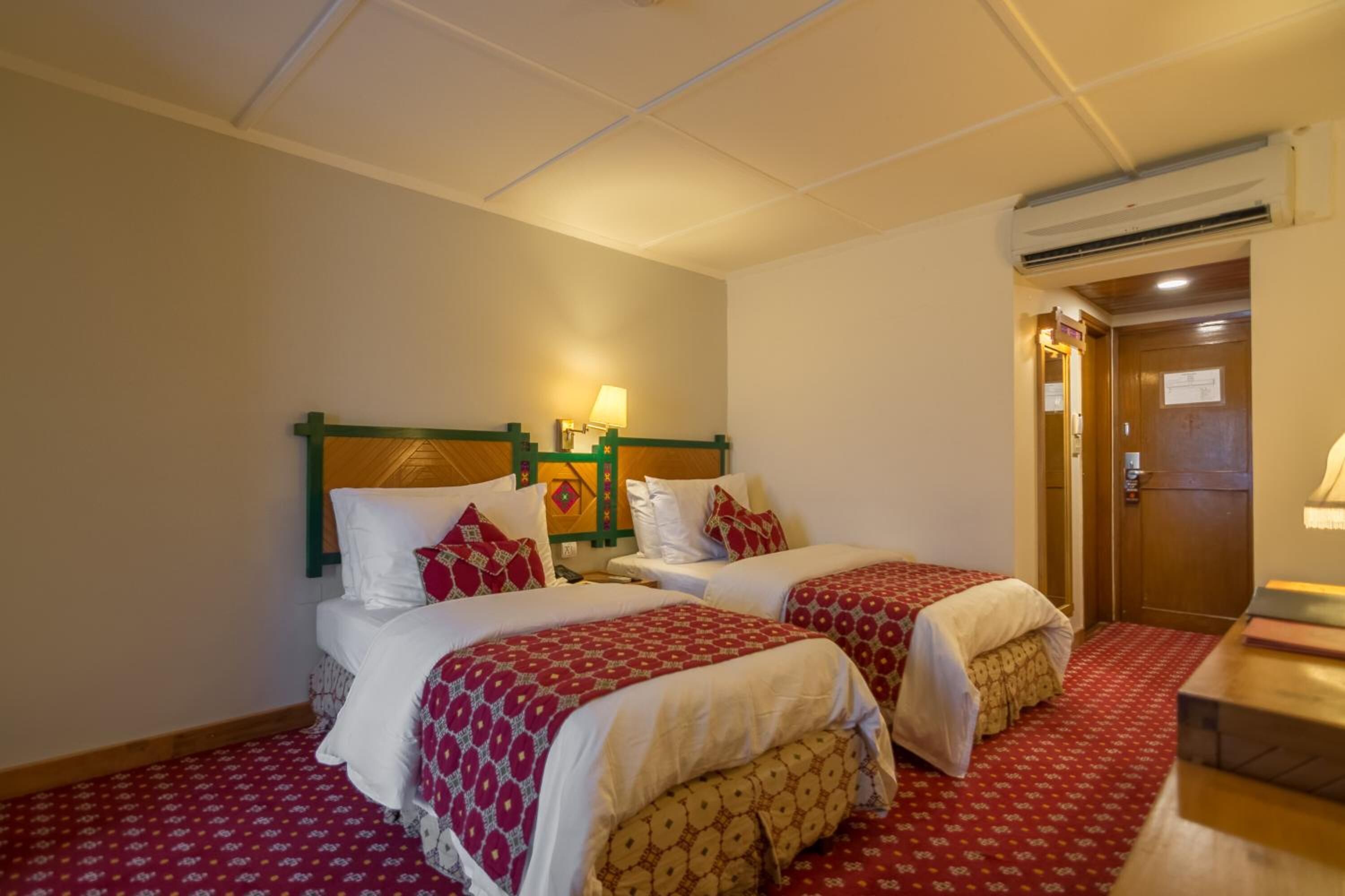 A hotel room with two beds, a wooden headboard, a nightstand, a lamp, and a wooden desk.
