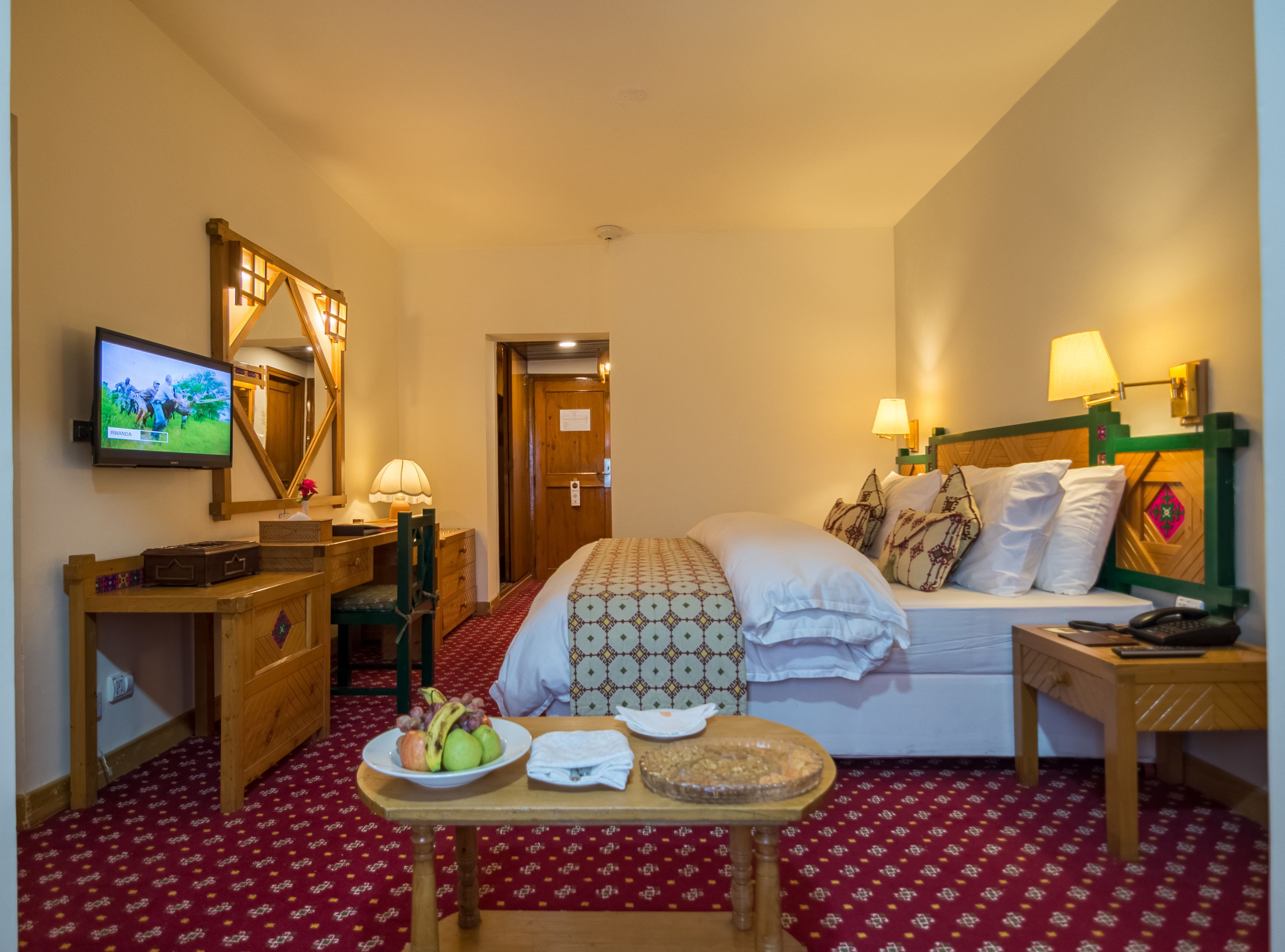 A hotel room with a bed, bedside table, lamp, television, and a small table with fruit and a plate.