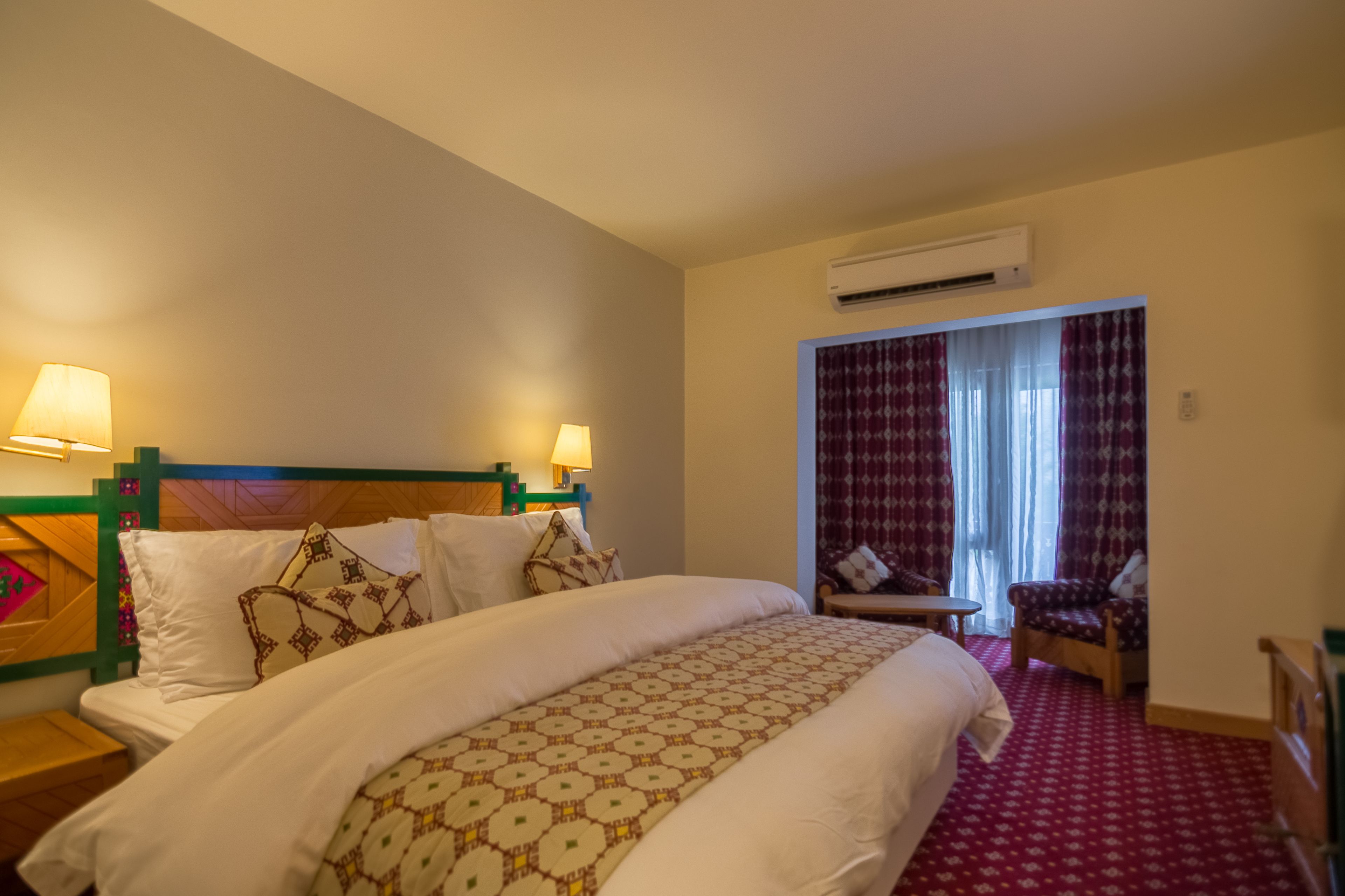 A hotel room with a large bed, two bedside lamps, a small table, a chair, and a window with curtains.