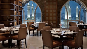 Un restaurante con mesas dispuestas para comer, amplios ventanales arqueados y vistas a una mezquita con minaretes.