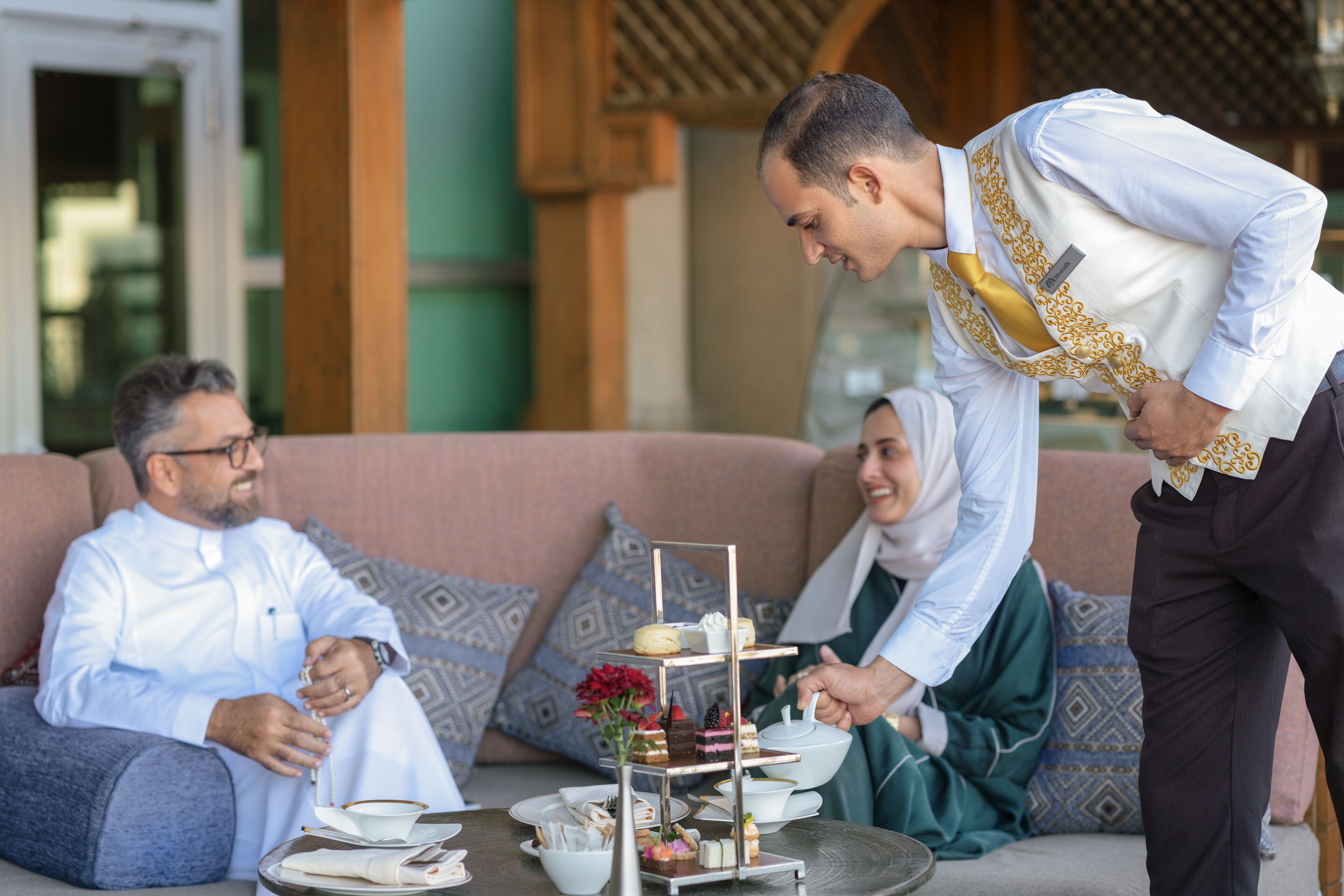 A waiter serving tea and pastries to guests seated on a sofa.