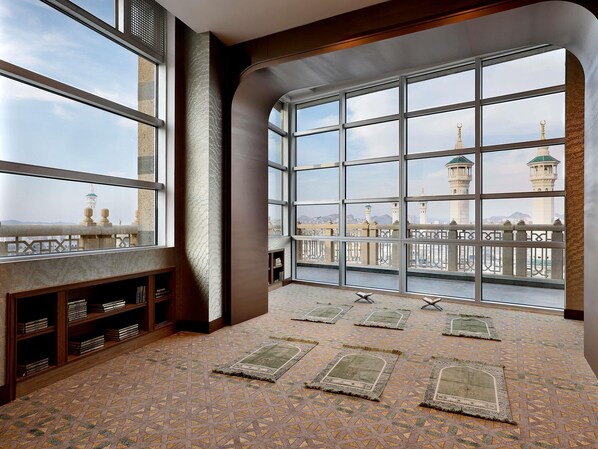 A room with large windows, a bookshelf, and prayer rugs on the floor.