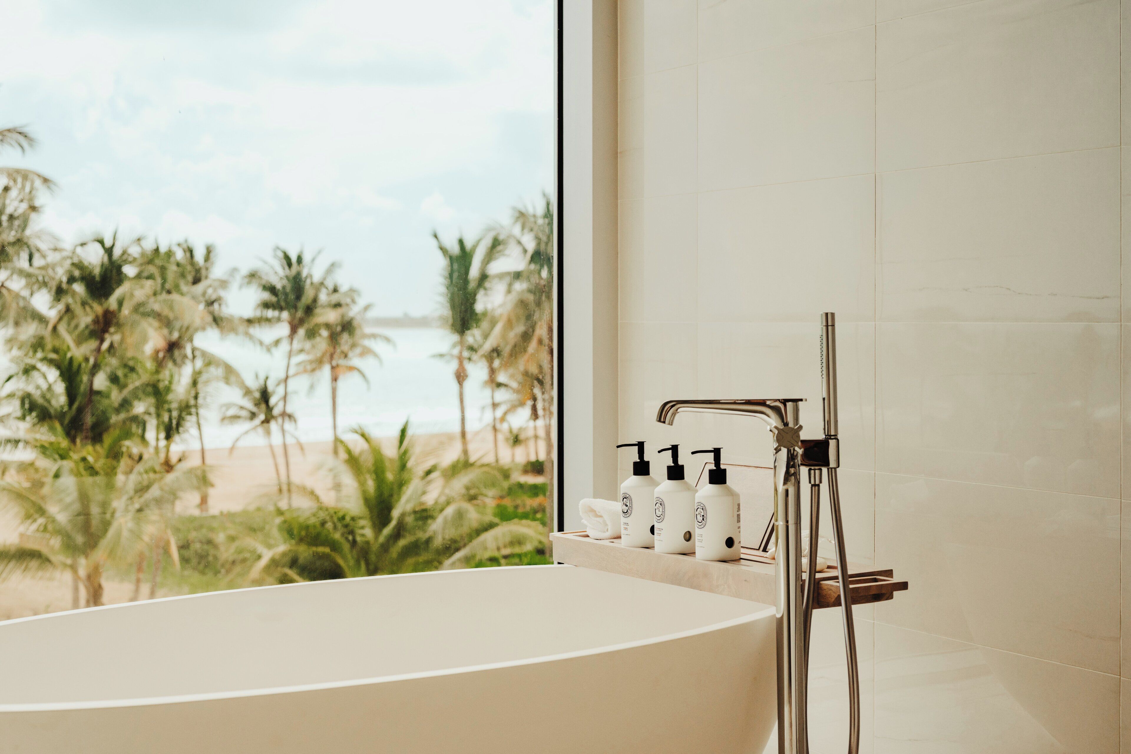 Un baño con bañera, ducha y ventana con vista a palmeras y una playa.