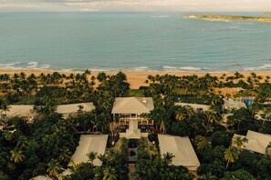 An aerial view of a resort with a central building surrounded by palm trees, a beach, and a pool.