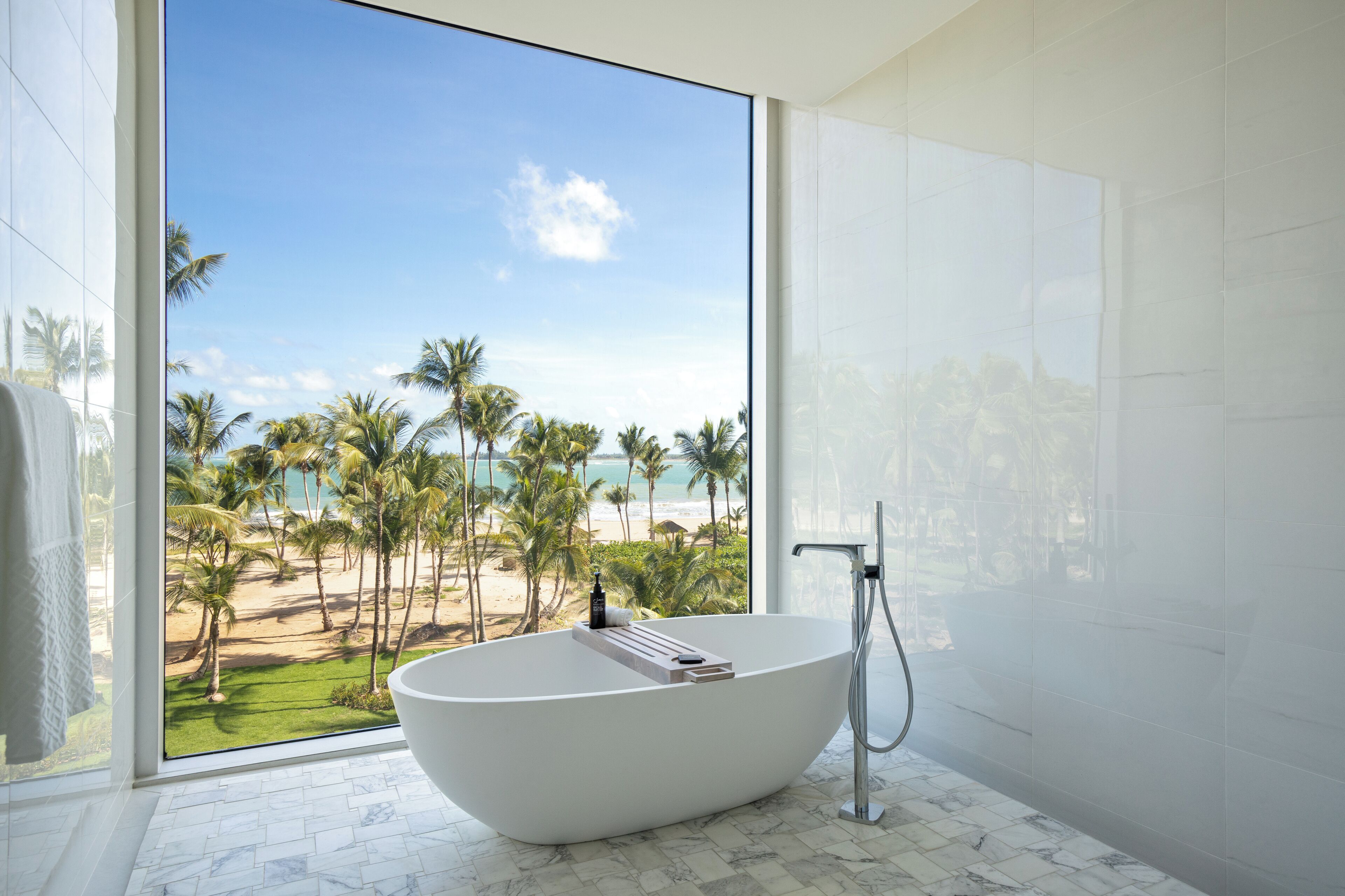 Un baño moderno con una bañera independiente, un ventanal con vistas a una playa y palmeras, y piso de mármol.
