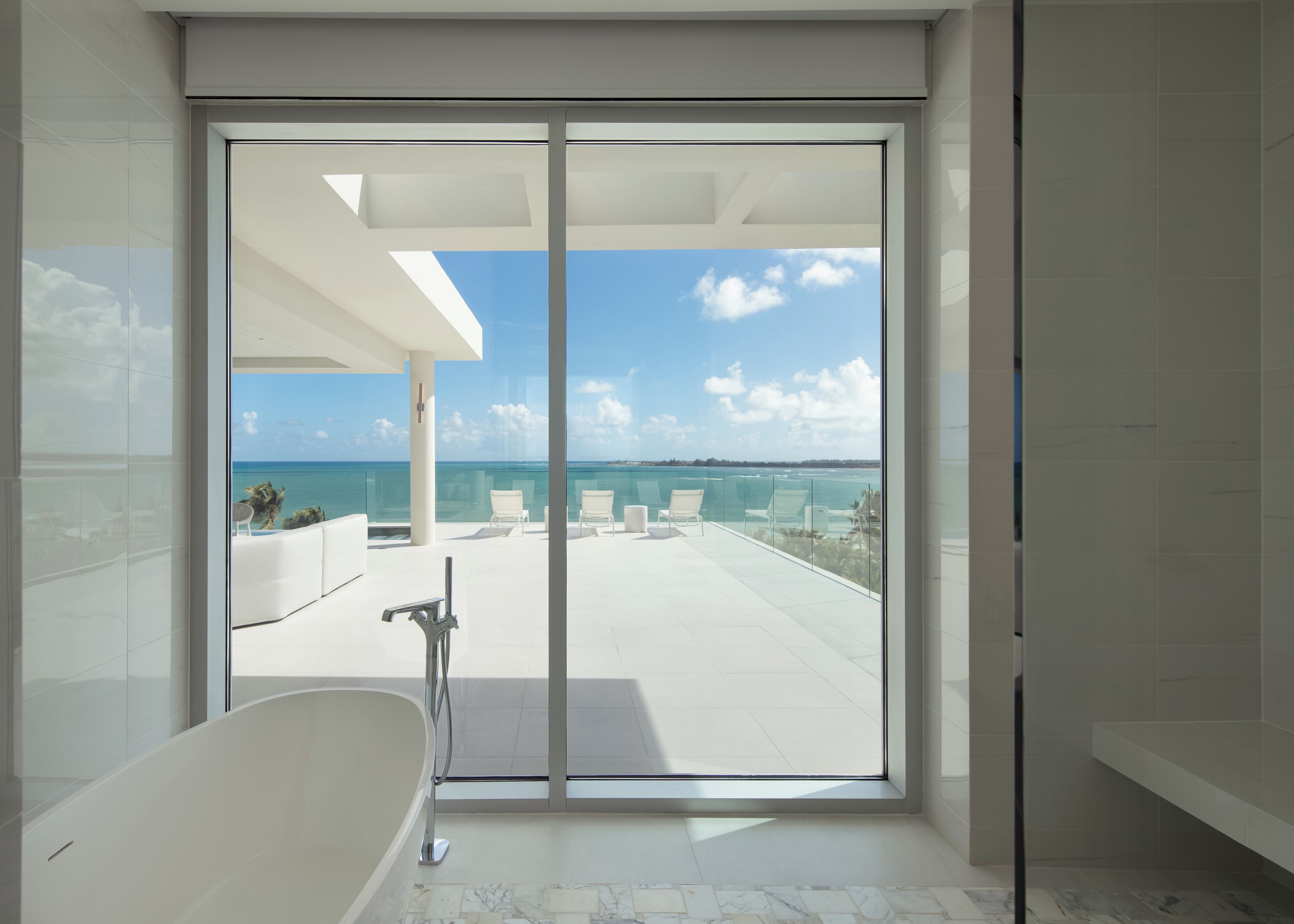 Un baño moderno con una bañera grande, un balcón con cerramiento de vidrio y vista al océano, y un cielo despejado.