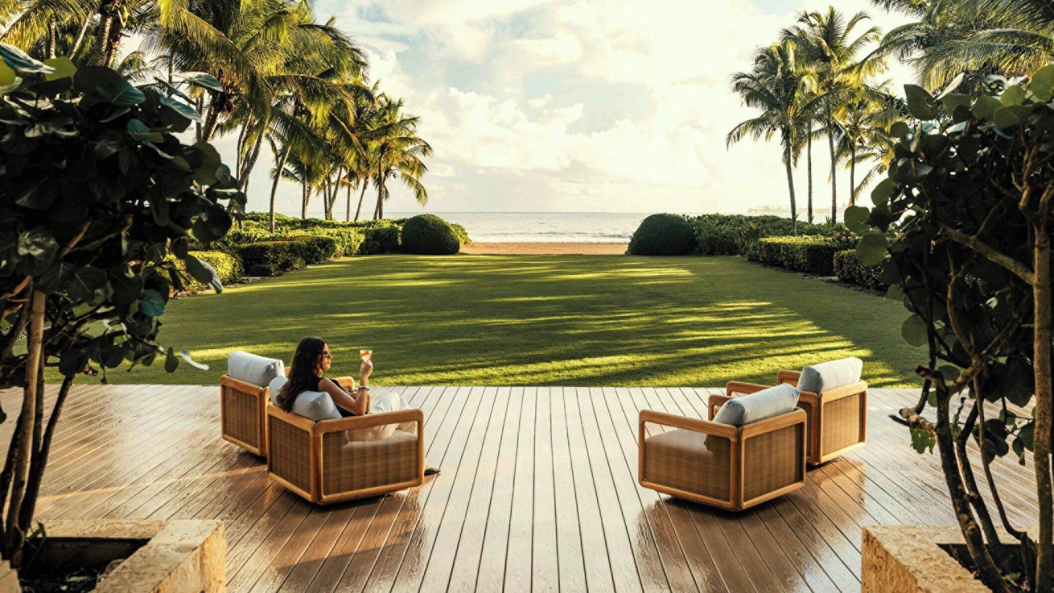 A deck with two wicker chairs facing a beach with palm trees and a clear sky.