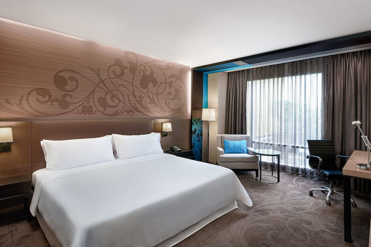 A hotel room with a large bed, a desk with a chair, a lamp, and a view of the outdoors through large windows.