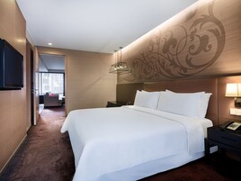 A hotel room with a large bed, a flat-screen TV mounted on the wall, a bedside table with a lamp, and a decorative wall panel.