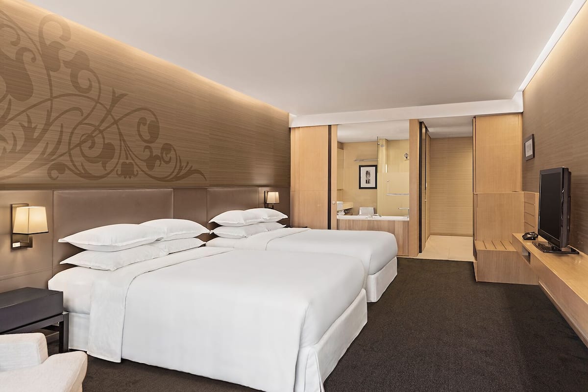 A hotel room with two beds, a desk, a TV, and a bathroom visible through an open door.