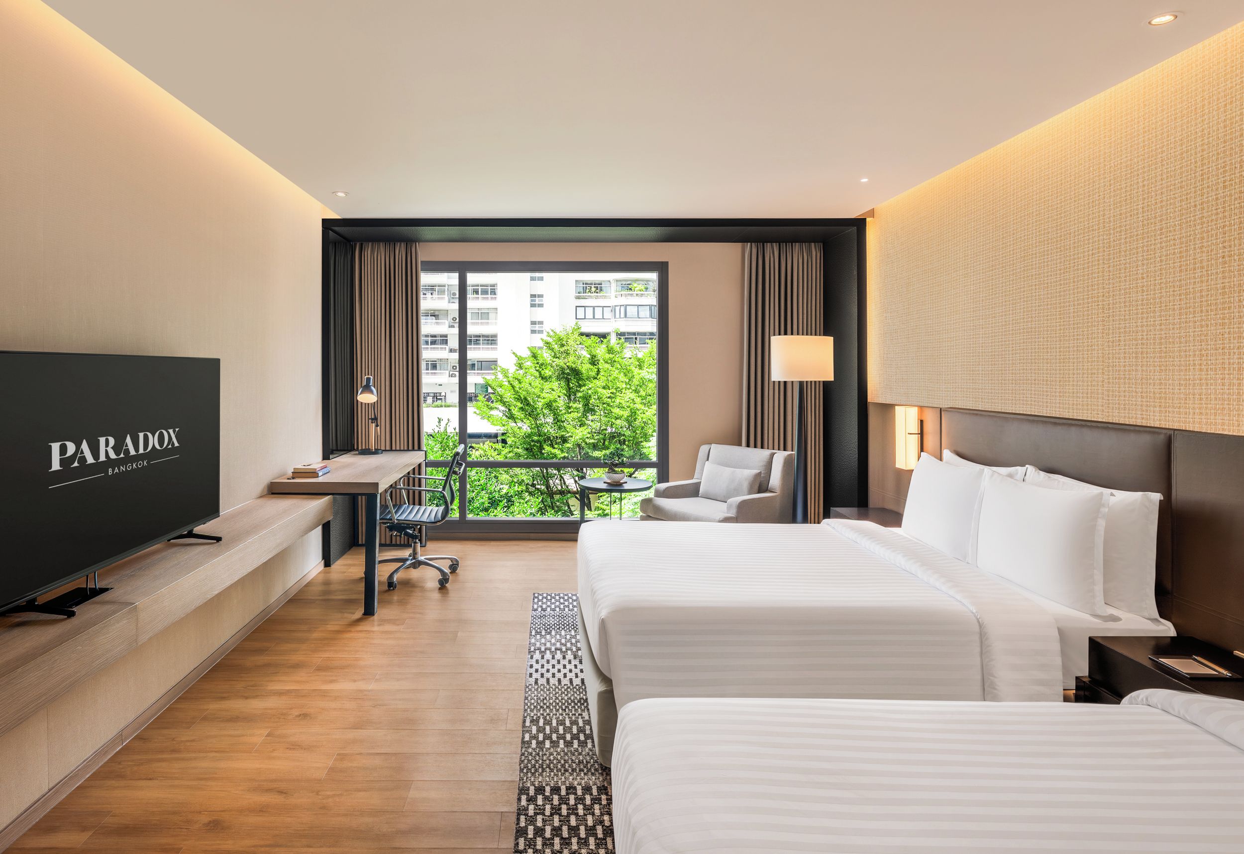 Une chambre d’hôtel avec un grand lit, un bureau, une chaise, une télévision et un balcon offrant une vue sur les bâtiments et la verdure.