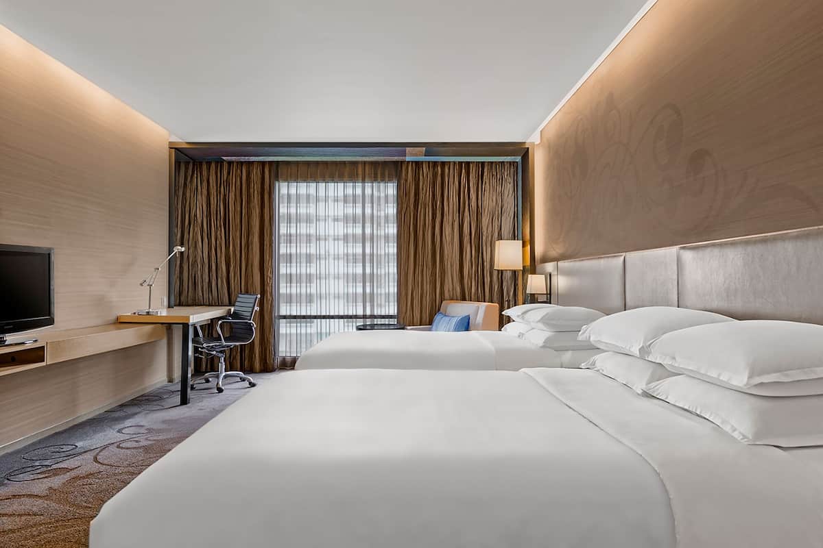 A hotel room with two beds, a desk with a TV, a chair, a lamp, and a window with curtains.