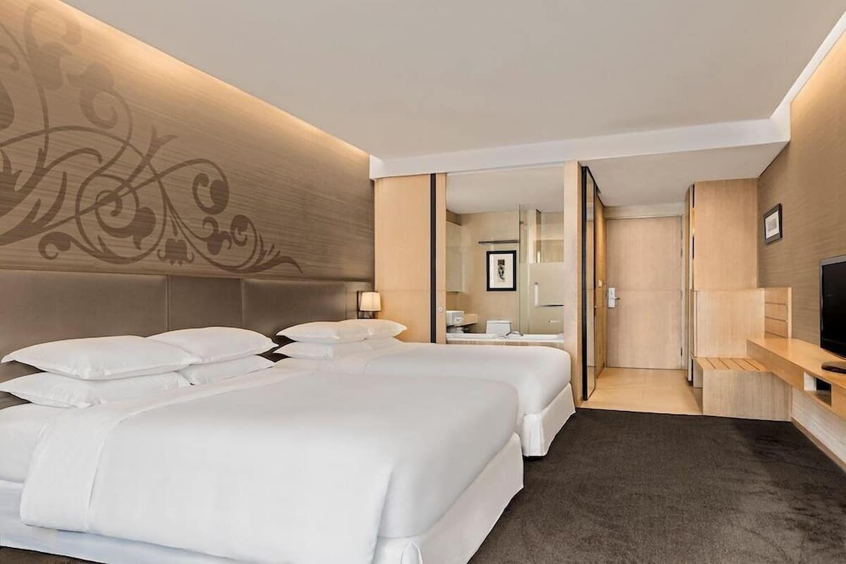 A hotel room with two beds, a TV, a desk, and a bathroom visible through a glass door.