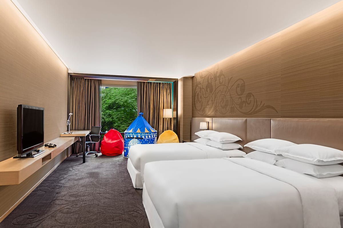A hotel room with two beds, a TV, a desk, and a view of greenery outside.