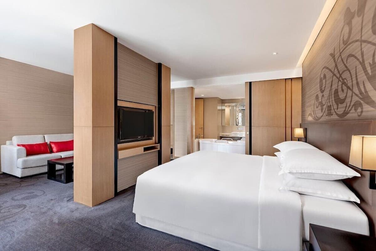 A modern hotel room with a large bed, a flat-screen TV, a sofa with red cushions, and a bathroom visible through a glass partition.