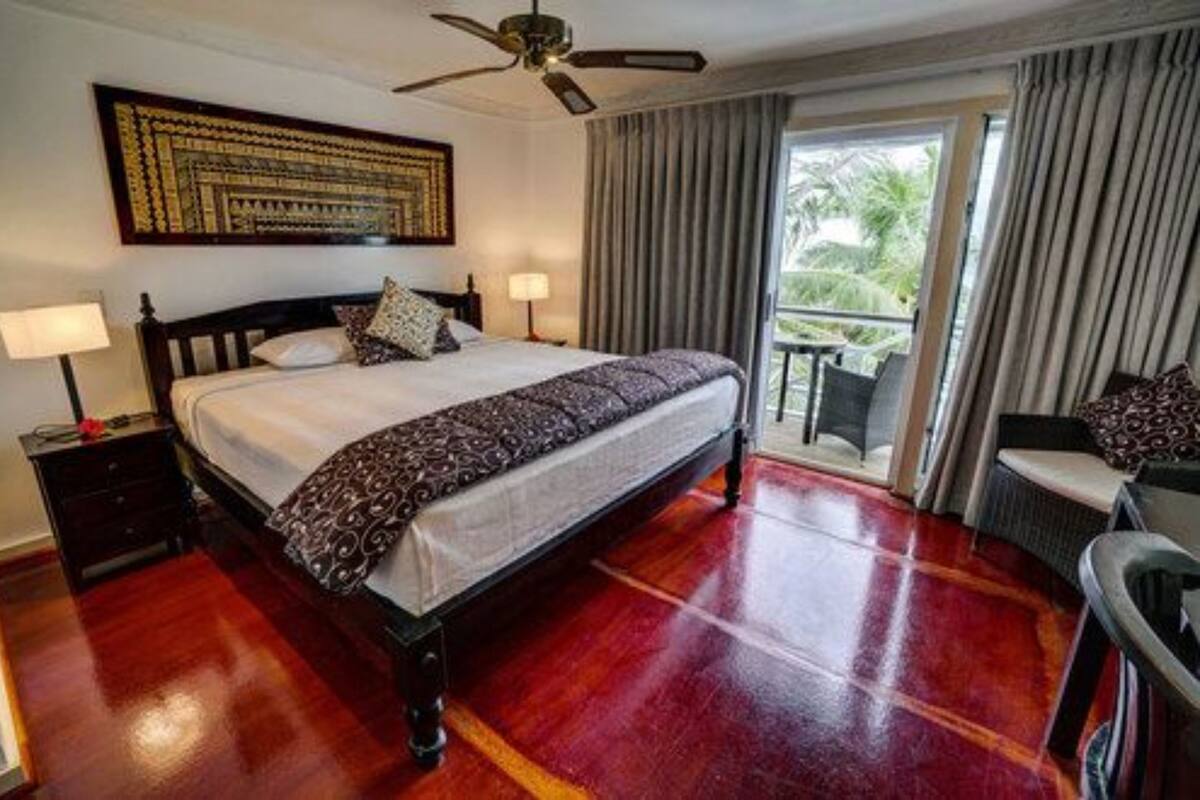 A hotel room with a large bed, a ceiling fan, a balcony with curtains, and a view of greenery outside.