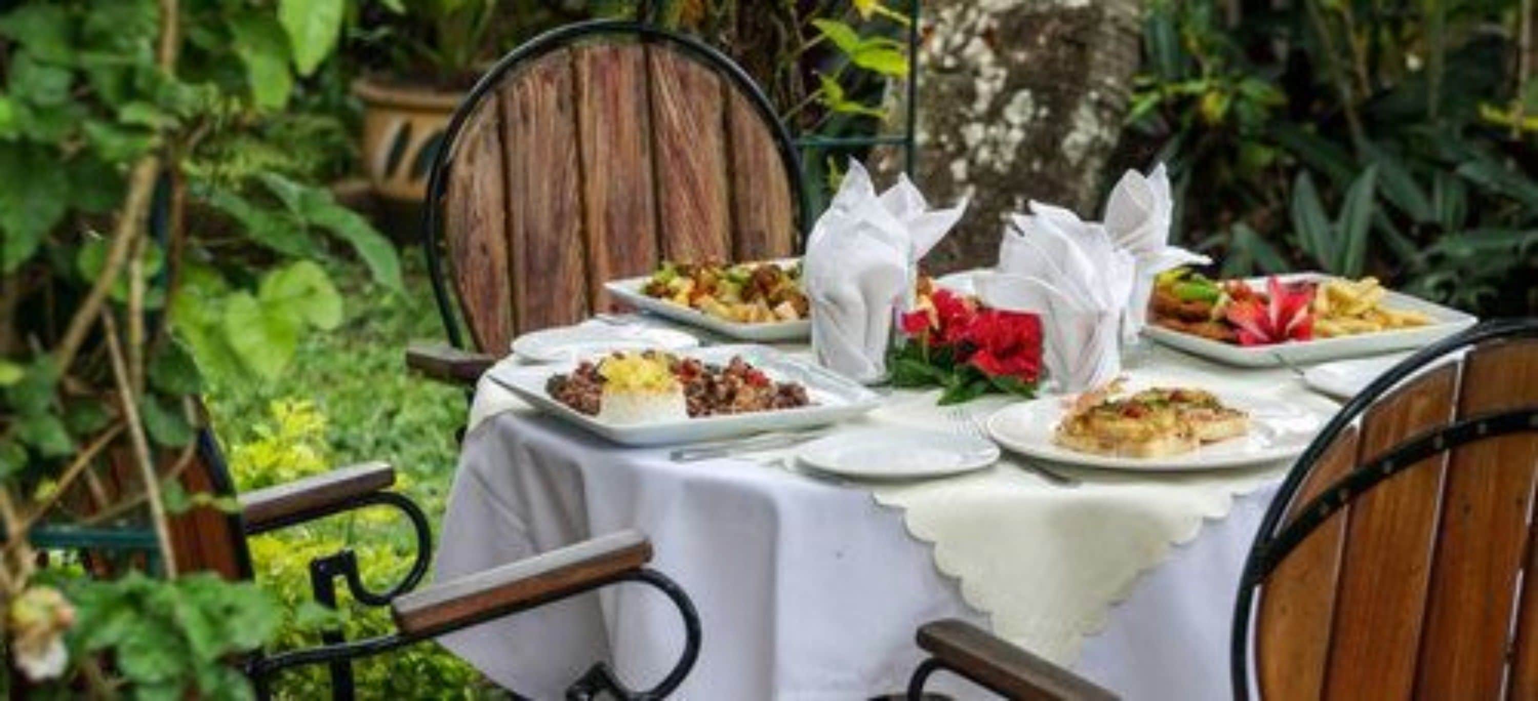 Mesa de jantar ao ar livre posta com pratos de comida, cercada por cadeiras e vegetação.