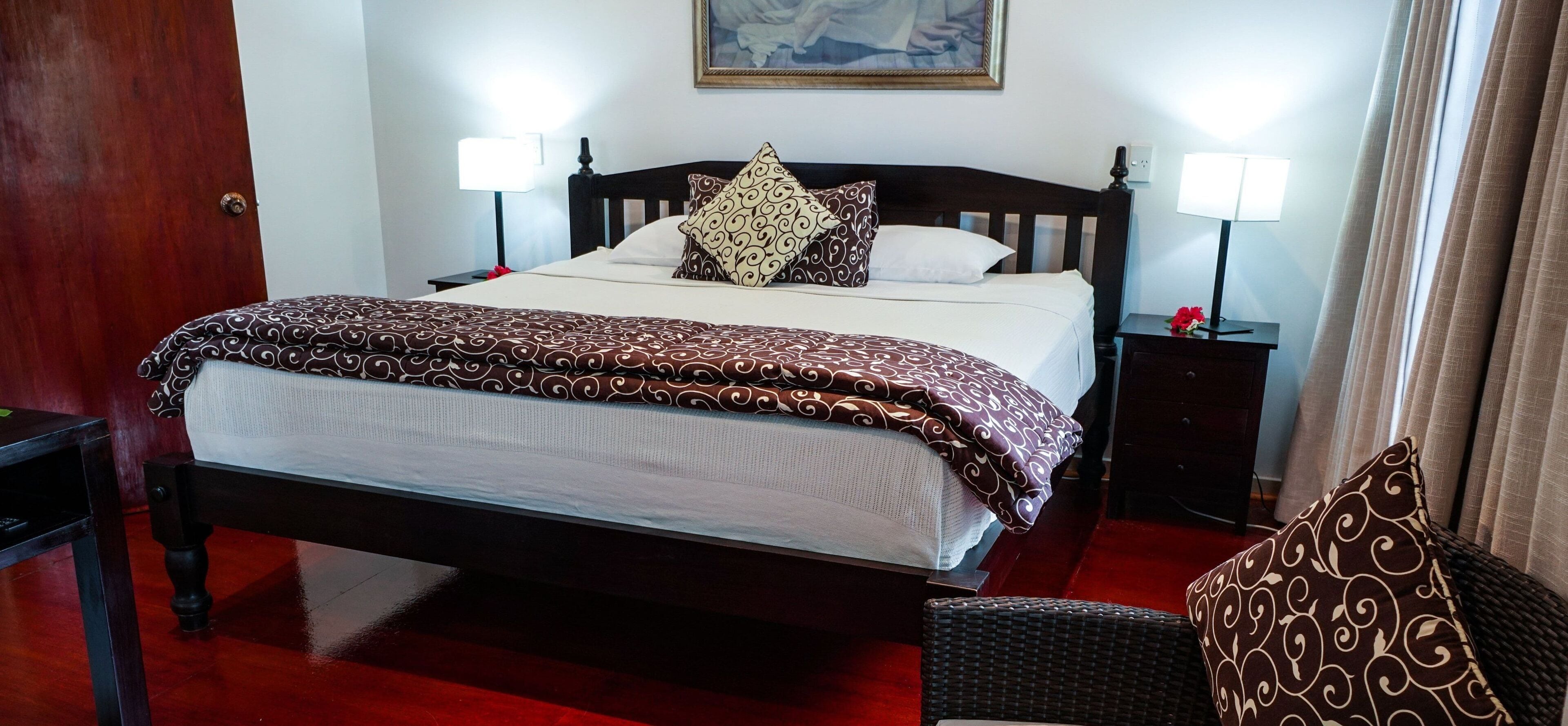 A neatly made bed with patterned bedding, two bedside lamps, a framed artwork above the bed, and a red carpeted floor.