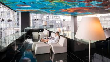 Un moderno vestíbulo de hotel con un mural en el techo, barandillas de vidrio y zonas de estar.
