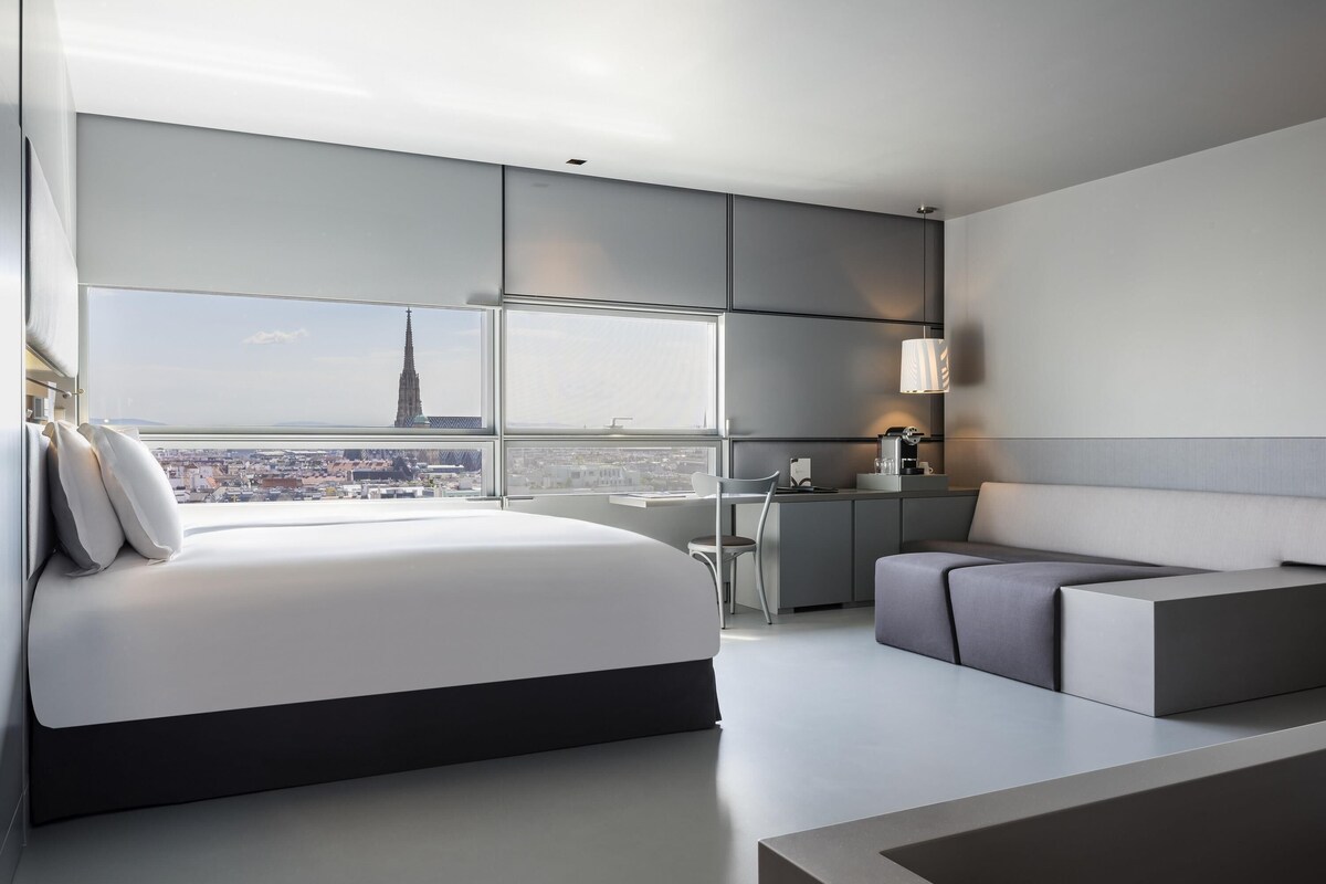 A modern hotel room with a large bed, a seating area, a desk, and a view of a cityscape.