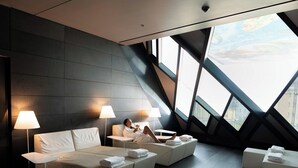 Una habitación de hotel moderna con una cama grande, dos mesitas de noche con lámparas, un sofá y vistas a la ciudad a través de amplios ventanales.