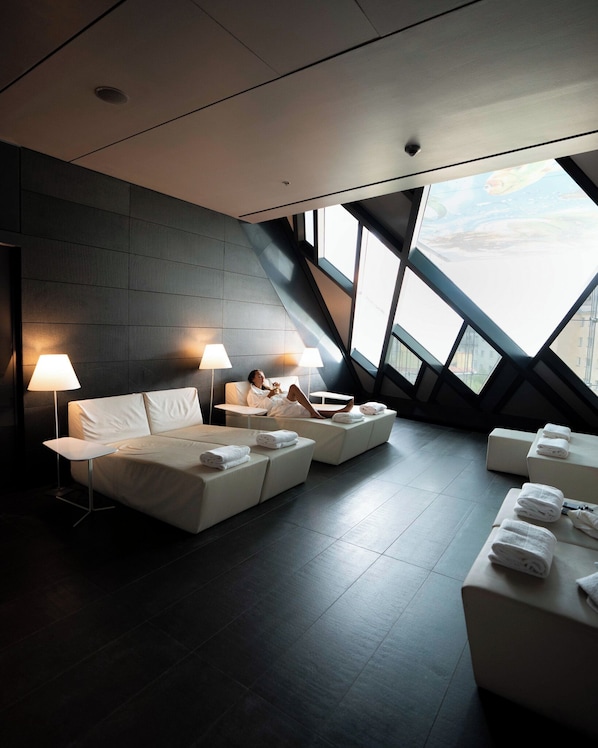 Ein modernes Hotelzimmer mit einem großen Bett, zwei Nachttischen mit Lampen, einem Sofa und Blick auf die Stadt durch große Fenster.