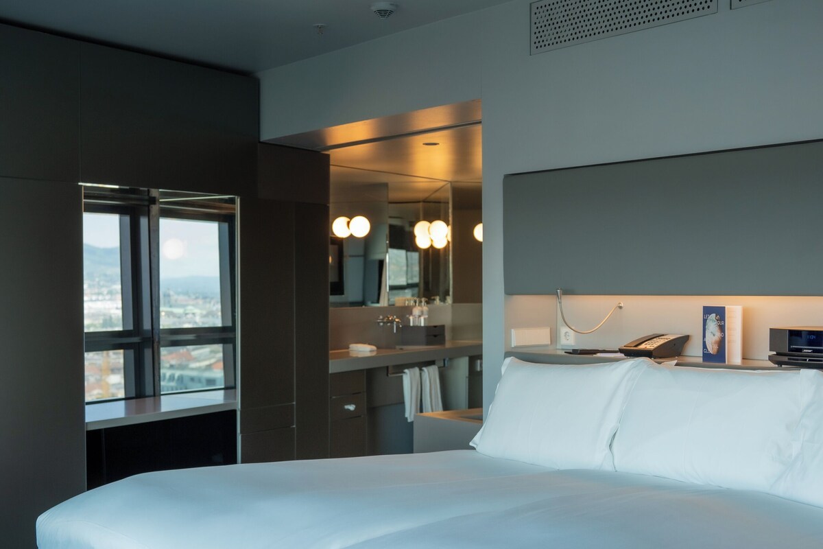 Una habitación de hotel moderna con una cama grande, un baño con espejo y un ventanal con vistas a la ciudad.