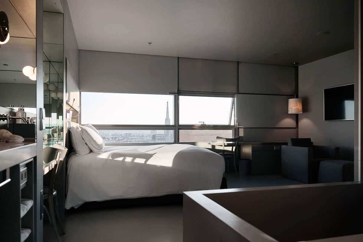 Ein modernes Hotelzimmer mit einem großen Bett, einem Schreibtisch, einem Sessel, einem Fernseher und Blick auf die Stadtlandschaft.